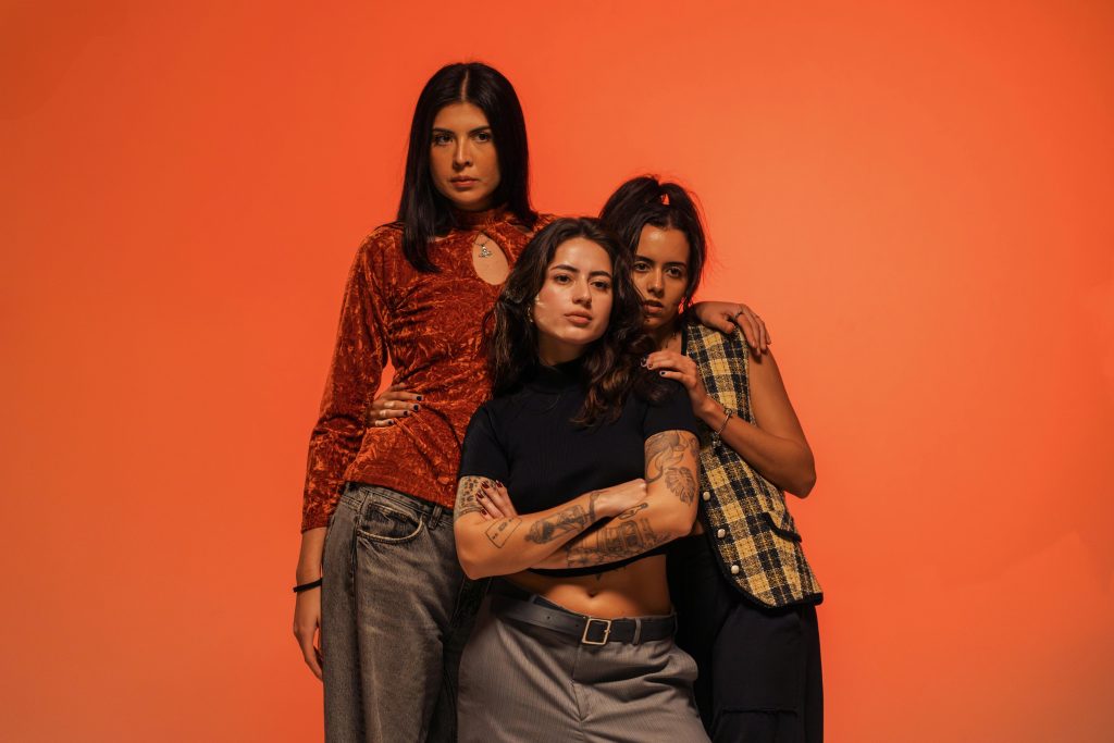 Three confident women pose against a vibrant orange backdrop, showcasing modern fashion and empowerment.