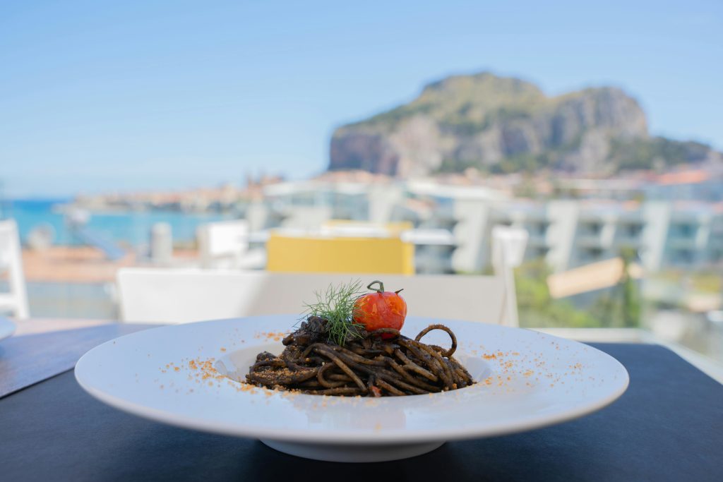 pexels-photo-34342500-34342500 Plate of squid ink pasta with tomato and herbs, set against a beautiful seaside view.